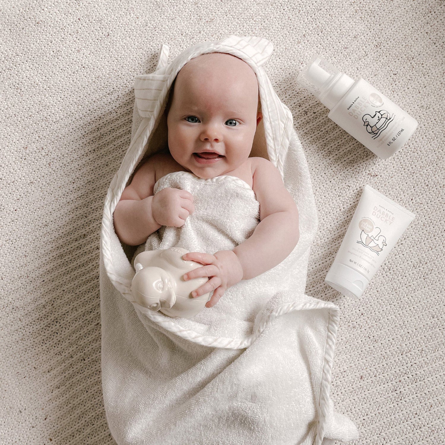 A smiling baby wrapped in a soft, cream-colored towel lies on a textured blanket, holding a toy. Nearby, Dabble & Dollop Fragrance Free Infant Lotion and Dabble & Dollop Fragrance Free Infant Wash are placed on the blanket.