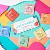 Colorful flat lay featuring the Just a Dabble Minis Set by Dabble & Dollop, surrounded by six vibrant travel-size packets: coconut, tangerine, honeydew, blueberry, strawberry, and lemon.