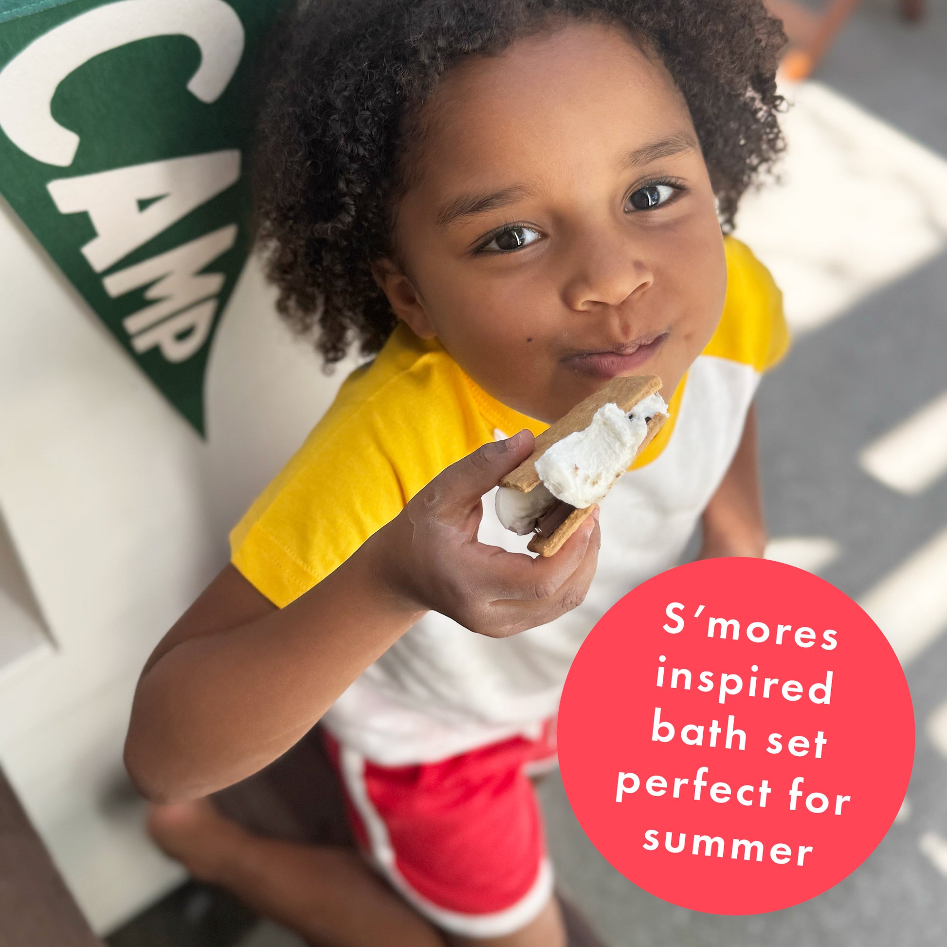 A smiling child in yellow and white with red shorts holds a s’more outside. Text on a pink circle reads: “S'more Bath Set by Dabble & Dollop—perfect for summer.” A green CAMP sign is visible in the background.