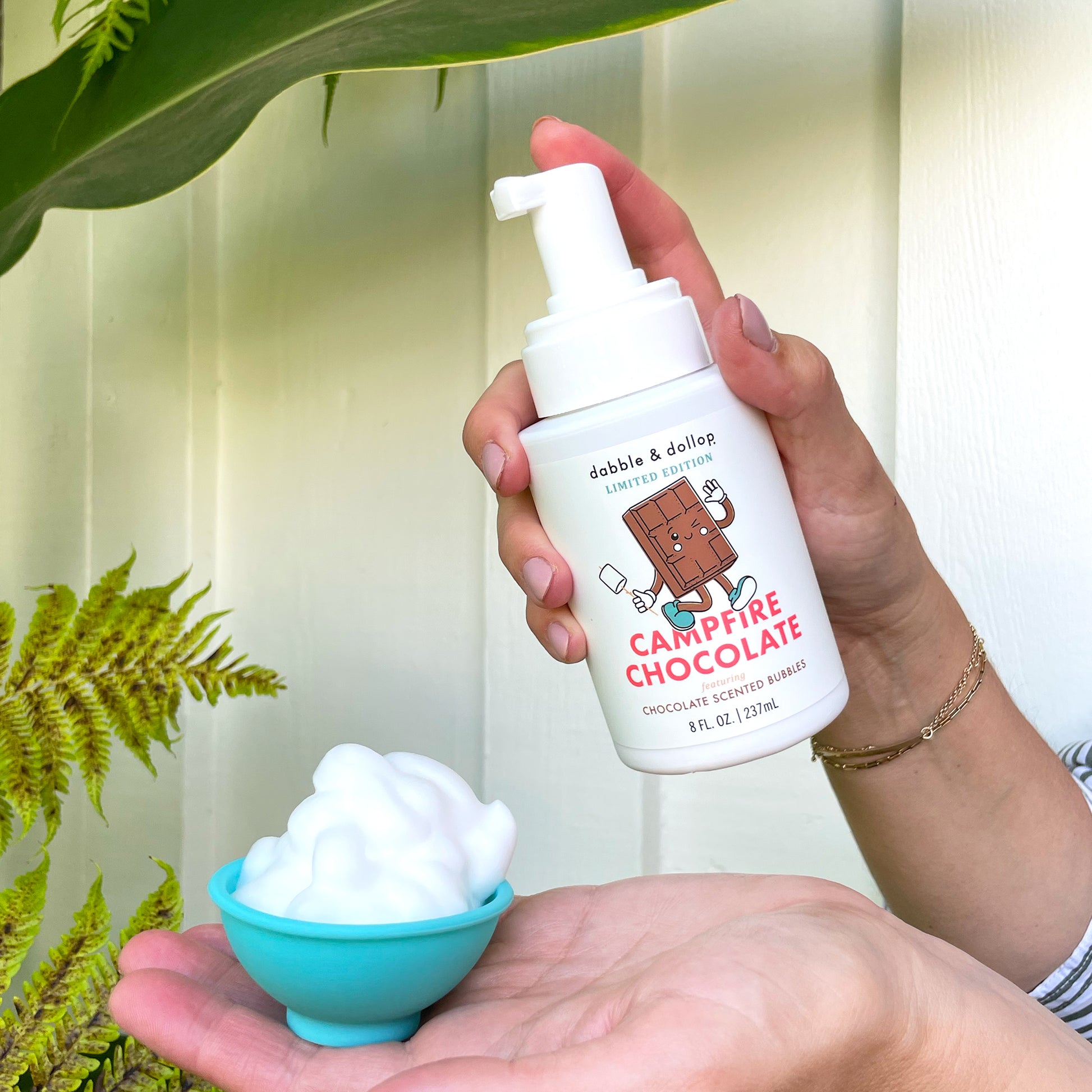 A hand holds a bottle of Dabble & Dollop Campfire Chocolate Whip, a chocolate-scented foaming soap ideal as a kids' body wash, while another hand displays a small blue bowl brimming with white foam. A green plant and light-colored wall are seen in the background.