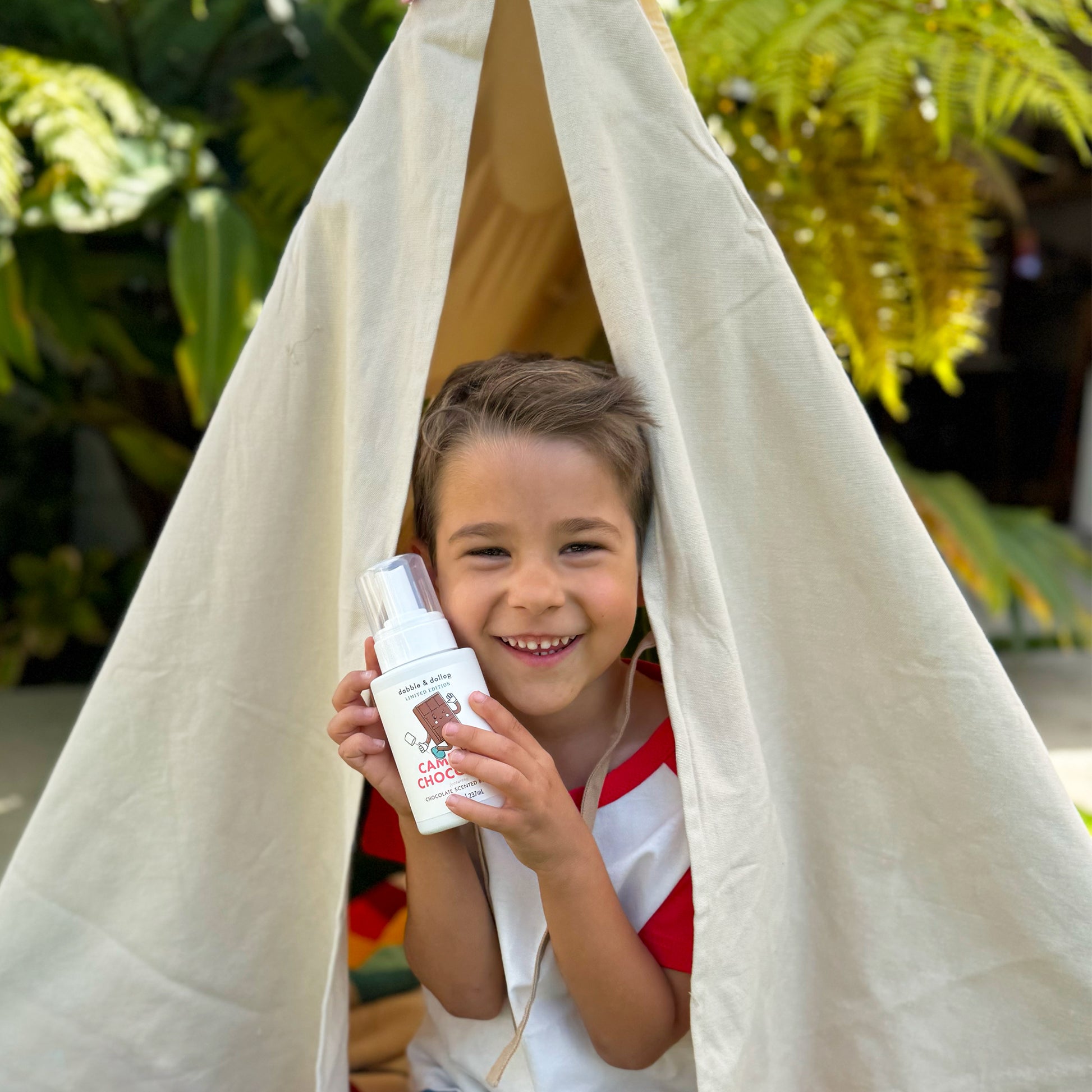 A smiling child sits inside a beige fabric tent outdoors, holding a bottle of Dabble & Dollop Campfire Chocolate Whip foaming kids body wash, with green plants in the background.