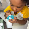 A young child in a yellow and white shirt stirs a white bottle with a teal lid, focusing intently. Nearby on the table in the kitchen, a tray with marshmallows awaits. The bottle contains Dabble & Dollop’s Campfire Chocolate Whip—an imaginative 3-in-1 shampoo, kids body wash, and bubble bath that turns bathtime into playtime fun.