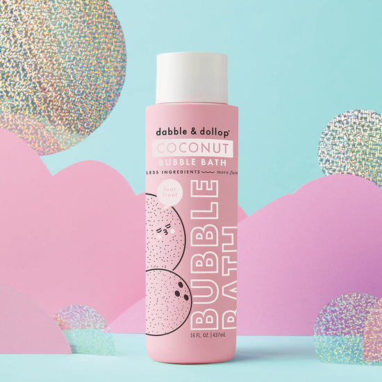 A bottle of Dabble & Dollop Coconut Bubble Bath, adorned with playful coconut illustrations on the label, sits on a pastel backdrop accented by shiny iridescent circles and pink cloud shapes. This gentle, hypoallergenic, and tear-free Coconut Bubble Bath brings fun to bath time for little ones.
