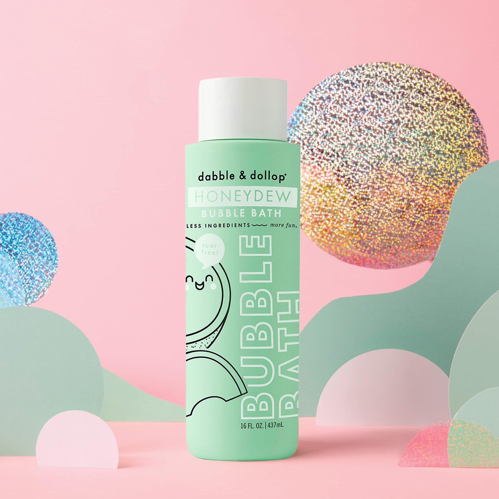 A green bottle of Dabble & Dollop Honeydew Bubble Bath is displayed among pastel shapes on a pink background, accented by shiny, iridescent decorative circles. Specially designed for kids, its tear-free formula is gentle on sensitive skin.