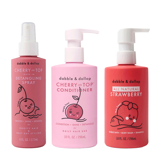 The Dabble & Dollop Bath to Brush Bundle includes three kids’ hair care bottles: a pink leave-in detangling spray, a light pink daily conditioner, and a red all-natural 3-in-1 bubble bath and shampoo with cherry and strawberry cartoon graphics.