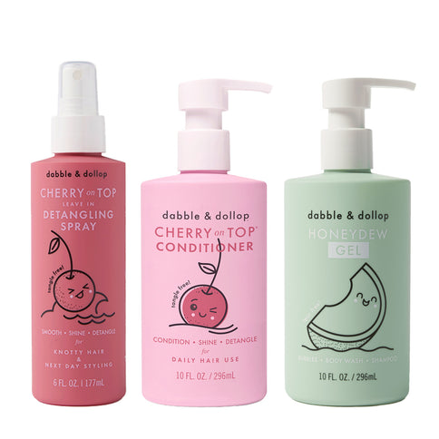 Three bottles of Dabble & Dollop hair and body products stand side by side: a Detangler, a Conditioner with cherry scent, and a honeydew gel—each featuring playful fruit illustrations on their labels.