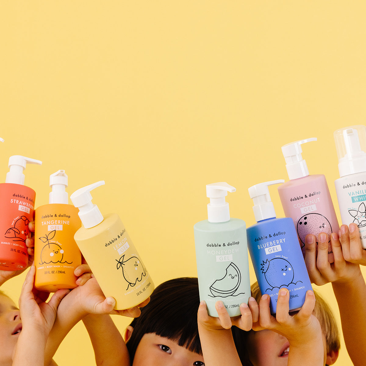 Children's hands hold up colorful Dabble & Dollop Build Your Own Bath Bundle bottles with cute fruit designs in scents like strawberry, tangerine, honeydew, and blueberry against a yellow background.