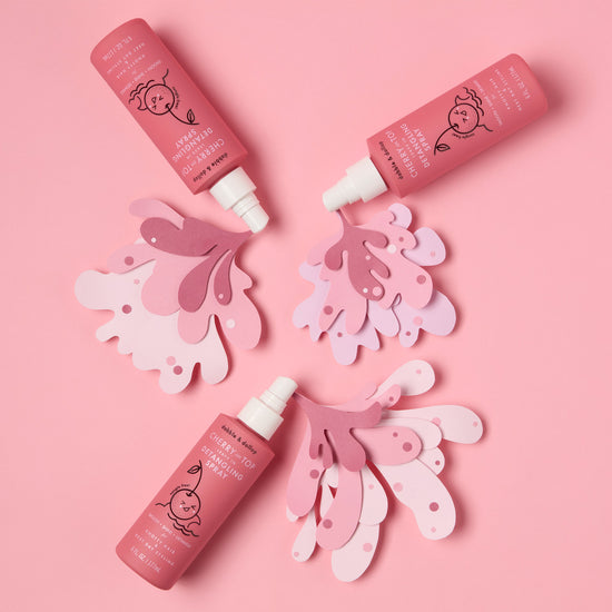 Four Cherry on Top Leave-In Spray Detangler bottles by Dabble & Dollop, with playful fruit art, are arranged on layered pink cutouts—ideal for gentle kids’ hair care or use as a detangling spray.