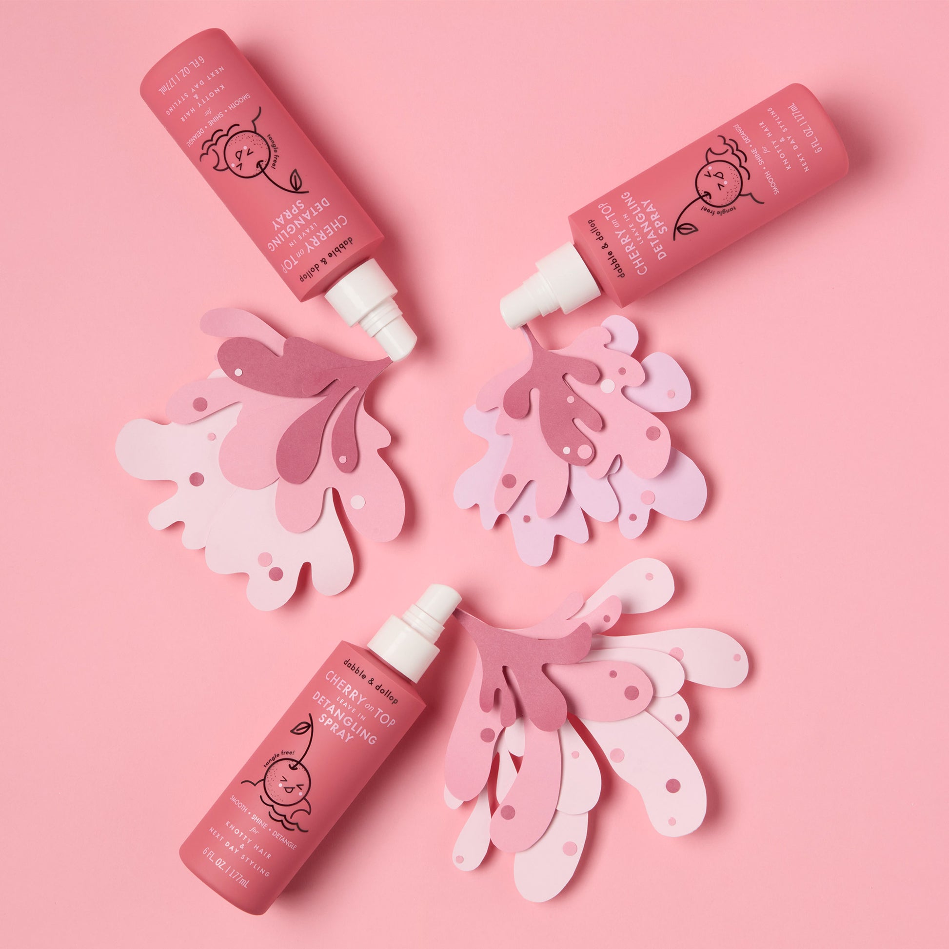 Four Cherry on Top Leave-In Spray Detangler bottles by Dabble & Dollop, with playful fruit art, are arranged on layered pink cutouts—ideal for gentle kids’ hair care or use as a detangling spray.