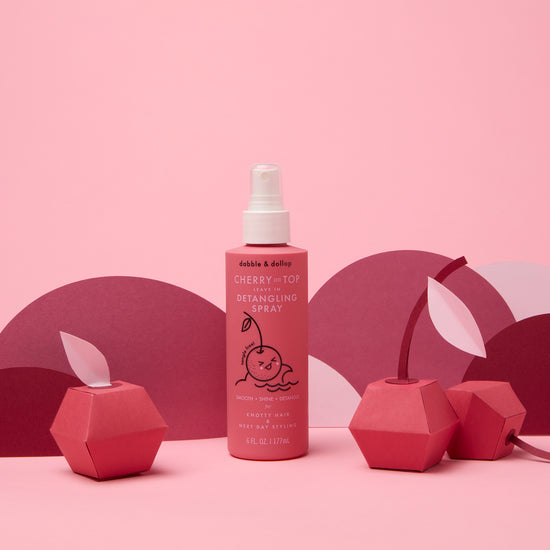 A bottle of Dabble & Dollop Cherry on Top Leave-In Spray Detangler stands on a pink surface, surrounded by geometric cherry paper decorations and set against a pink and purple background.