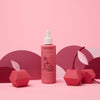A bottle of Dabble & Dollop Cherry on Top Leave-In Spray Detangler stands on a pink surface, surrounded by geometric cherry paper decorations and set against a pink and purple background.