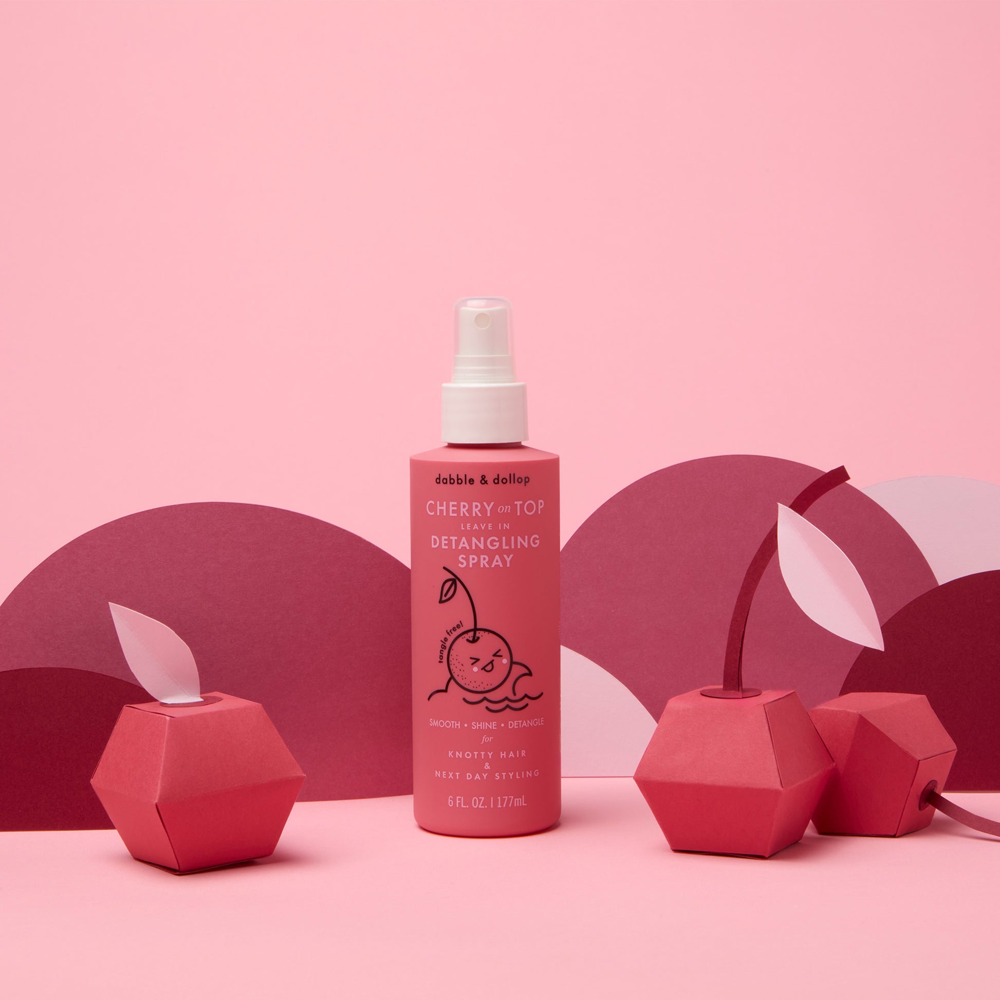 A bottle of Dabble & Dollop Cherry on Top Leave-In Spray Detangler stands on a pink surface, surrounded by geometric cherry paper decorations and set against a pink and purple background.