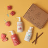 Flat lay of Dabble & Dollops Cherry Berry Conditioner & Bath Bundle on vibrant yellow: features a fruit-illustrated box, bottles labeled Strawberry and Cherry on Top Conditioner, a vanilla foaming bottle, cinnamon sticks, and paper strawberries.
