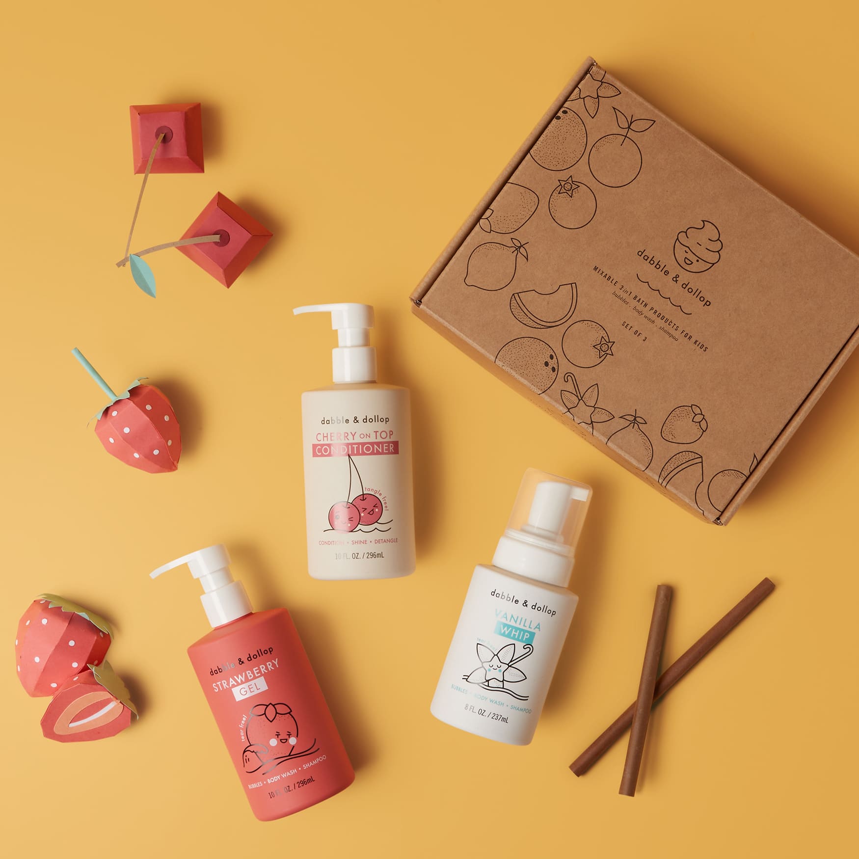 Flat lay of Dabble & Dollops Cherry Berry Conditioner & Bath Bundle on vibrant yellow: features a fruit-illustrated box, bottles labeled Strawberry and Cherry on Top Conditioner, a vanilla foaming bottle, cinnamon sticks, and paper strawberries.