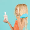 A young girl with long blonde hair, wearing an orange top, holds a Cherry Berry Conditioner & Bath Bundle by Dabble & Dollop in one hand and a large blue comb in the other, standing against a light turquoise background.