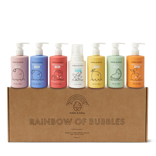Seven playful pastel bottles of Dabble & Dollop Rainbow of Bubbles Bath Bundle sit atop a cardboard box, featuring scents like blueberry, strawberry, and vanilla. These colorful bottles promise a delightful bubble bath or gentle body wash with cute fruit illustrations.