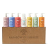 Seven playful pastel bottles of Dabble & Dollop Rainbow of Bubbles Bath Bundle sit atop a cardboard box, featuring scents like blueberry, strawberry, and vanilla. These colorful bottles promise a delightful bubble bath or gentle body wash with cute fruit illustrations.
