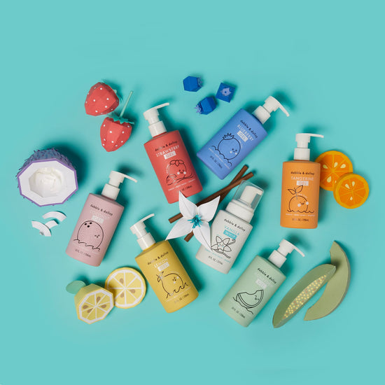 The Rainbow of Bubbles Bath Bundle by Dabble & Dollop features colorful shampoo and body wash bottles with playful animal illustrations on a teal background, surrounded by vibrant paper art fruits like strawberries, oranges, lemons, and coconuts.
