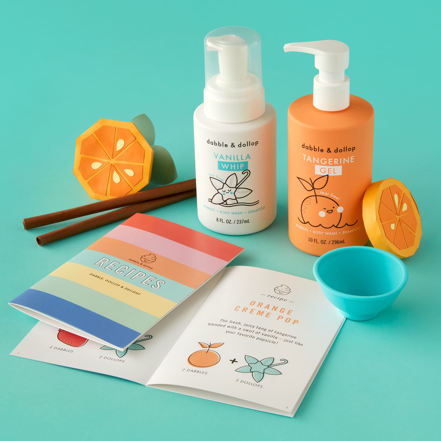 Bottles of Vanilla Whip and Tangerine Gel foam bath from Dabble & Dollop rest on a turquoise surface, surrounded by orange slice bath toys, a turquoise cup, and the vibrant Dabble & Dollop Recipe Booklet that inspires creativity and fun in your bath routine.