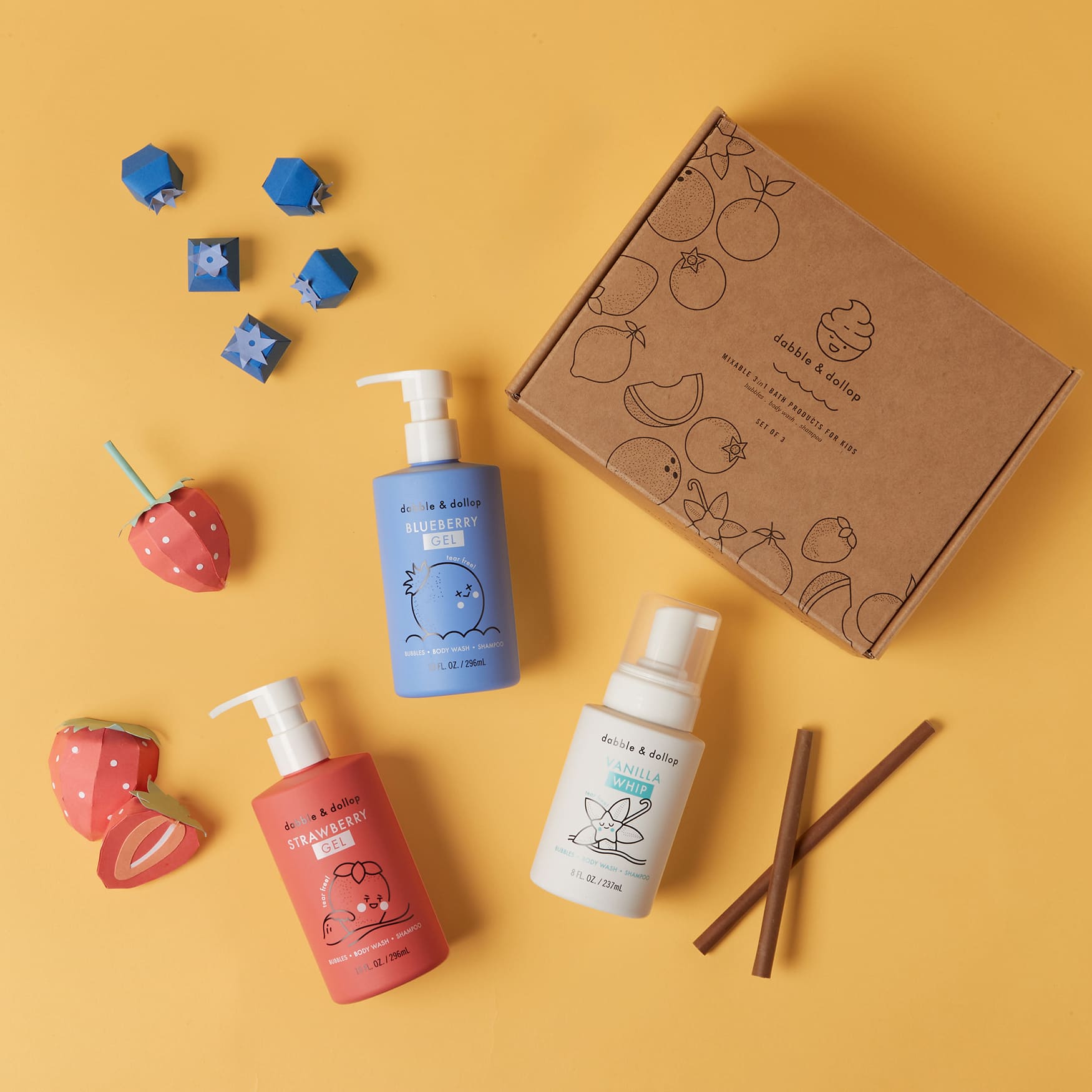 A flat lay on a yellow background features the Americana Bath Bundle from Dabble & Dollop, showcasing a fruit-illustrated box, two pump bottles labeled as strawberry and blueberry scents, and decorative strawberries and cinnamon sticks for a Strawberry Vanilla Blueberry Gels experience.