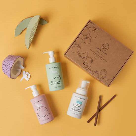 A Day at the Beach Bath Bundle by Dabble & Dollop, featuring a pink, green, and white bottle set against a yellow backdrop. A brown box with fruit illustrations sits nearby with cinnamon sticks and paper fruit decorations, highlighting its natural vegan cruelty-free essence.