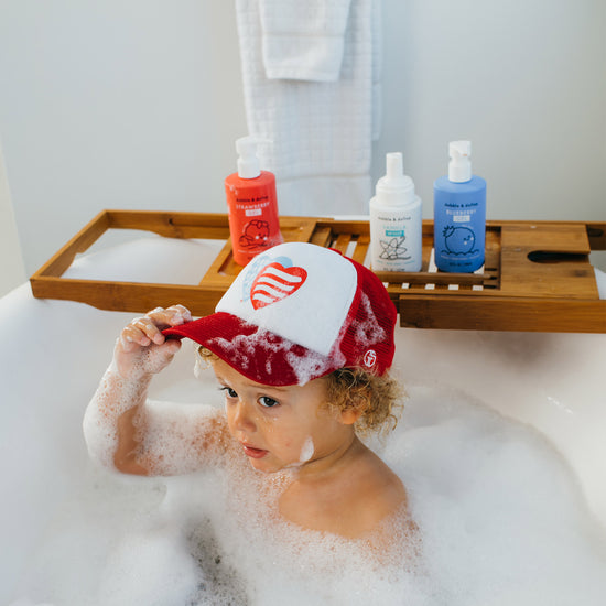 A young child with curly hair sits in a bubble bath, wearing a red and white heart cap. Nearby, Dabble & Dollops Americana Bath Bundle soap bottles and towel rest on a wooden bath tray.