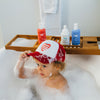 A young child with curly hair sits in a bubble bath, wearing a red and white heart cap. Nearby, Dabble & Dollops Americana Bath Bundle soap bottles and towel rest on a wooden bath tray.