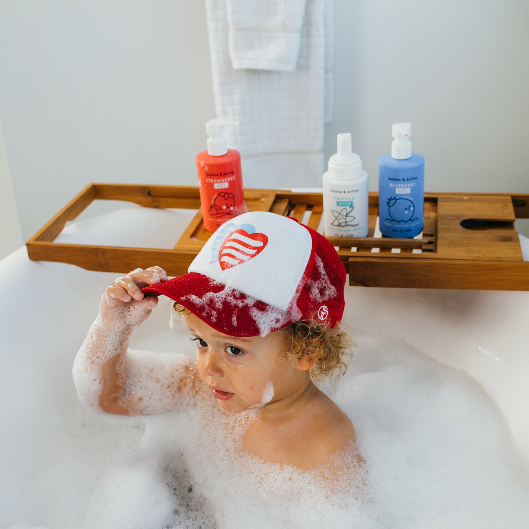 A young child with curly hair sits in a bubble bath, wearing a red and white heart cap. Nearby, Dabble & Dollops Americana Bath Bundle soap bottles and towel rest on a wooden bath tray.