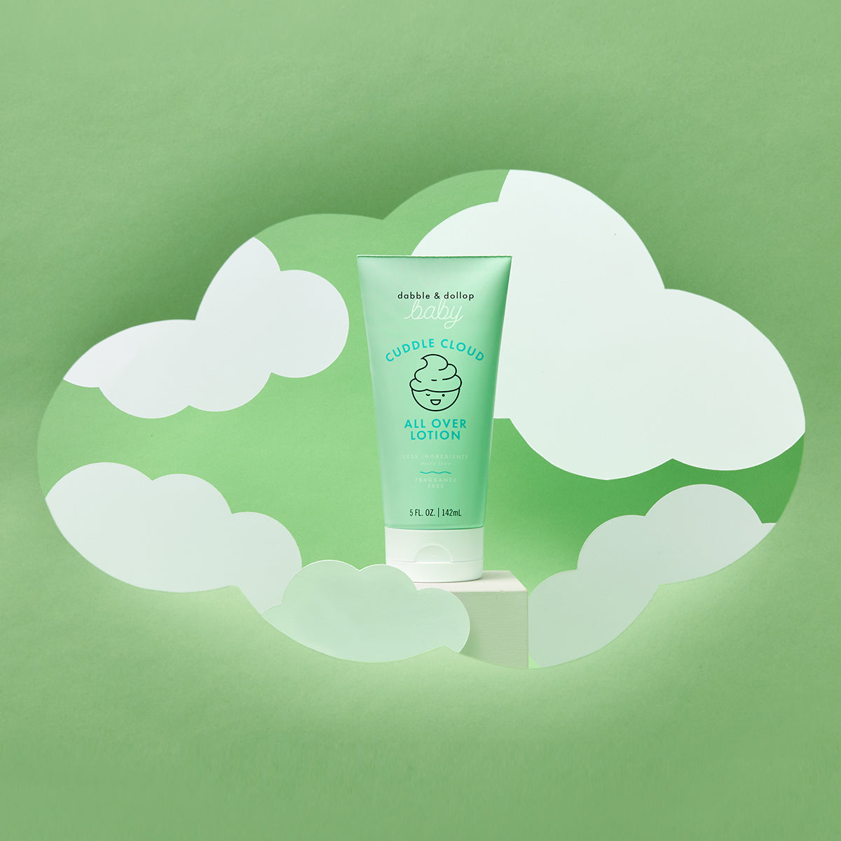 A tube of Dabble & Dollop Cuddle Cloud™ All Over Lotion stands on a white platform, surrounded by paper cut-out clouds with a green backdrop.