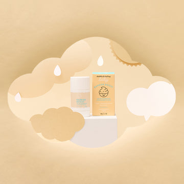 Dabble & Dollop’s Barrier Boost™ Skin Barrier Soothing Stick and its box, adorned with a smiling baby face logo, are showcased on a white pedestal with soft beige cloud, raindrop, and sun cutouts, emphasizing gentle skin barrier care.