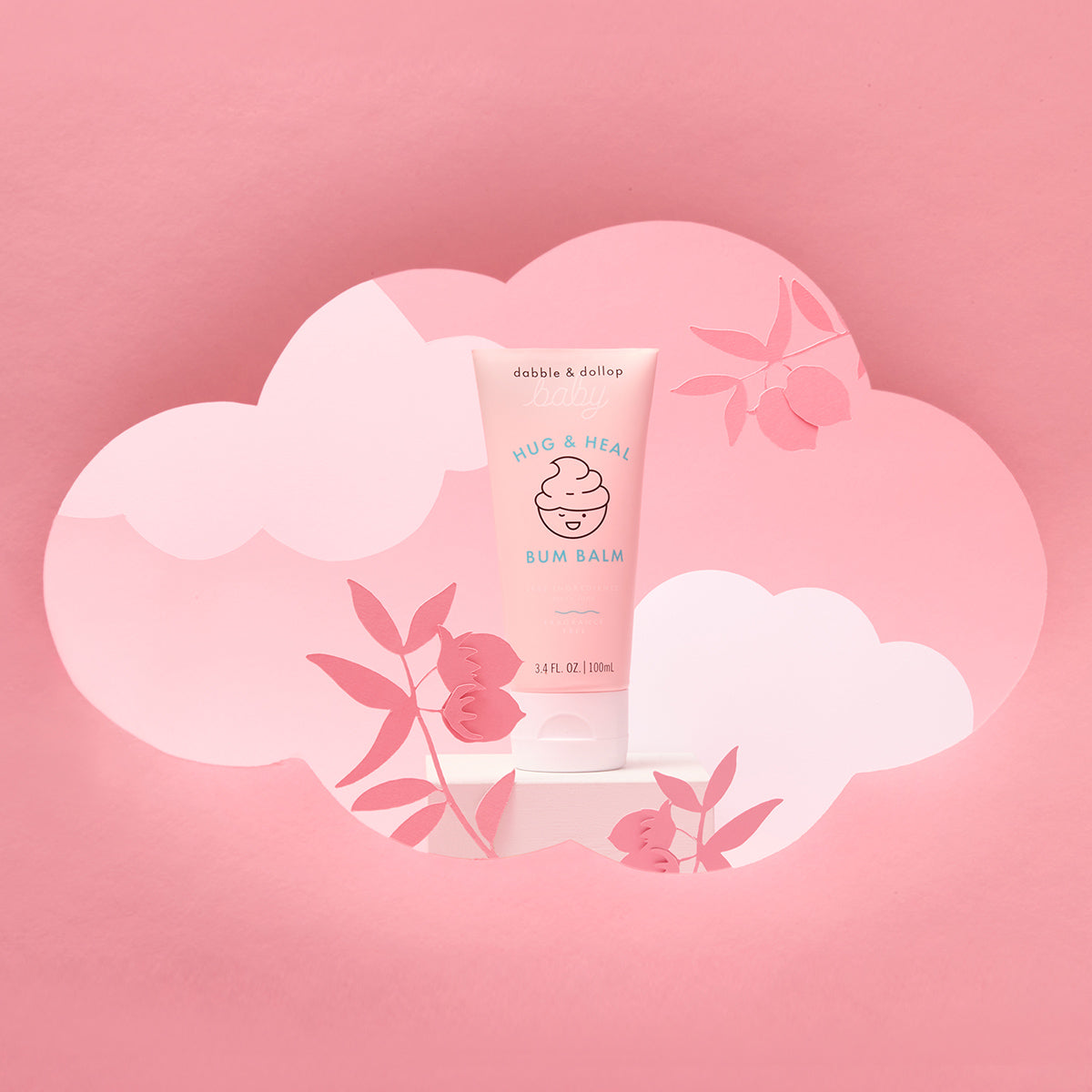 A tube of Dabble & Dollop Hug & Heal™ Bum Balm stands before a pink, cloud-shaped cutout and paper flowers—ideal for soothing and protecting your baby’s sensitive skin on a matching pink background.