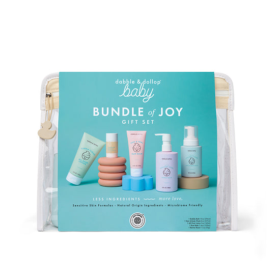 A clear zippered pouch displays the Dabble & Dollop Bundle of Joy Gift Set, featuring pastel-colored bottles and tubes of gentle skin care products, with a blue label listing contents and benefits.