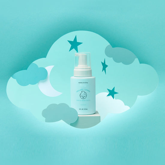 A white pump bottle labeled Lullaby Lather™ Hair & Body Wash by Dabble & Dollop stands before a blue background decorated with paper clouds, stars, and a crescent moon for a dreamy nighttime scene.
