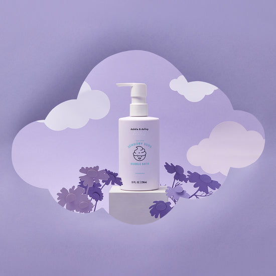 A bottle of Dabble & Dollop Sensory Suds™ Bubble Bath with a pump rests on a white block with cloud and flower cutouts against a purple background, creating a dreamy, whimsical scene.