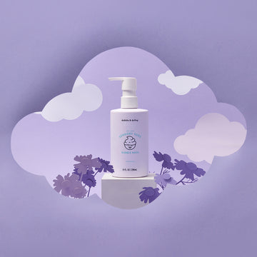 A bottle of Dabble & Dollop Sensory Suds™ Bubble Bath with a pump rests on a white block with cloud and flower cutouts against a purple background, creating a dreamy, whimsical scene.