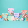 Four colorful bags of Dabble & Dollop Mini Bath Bombs are arranged among playful pastel props and shapes against a turquoise background. The packaging, reminiscent of kids’ bath bombs, features polka dots in various pastel colors for an extra burst of fun.