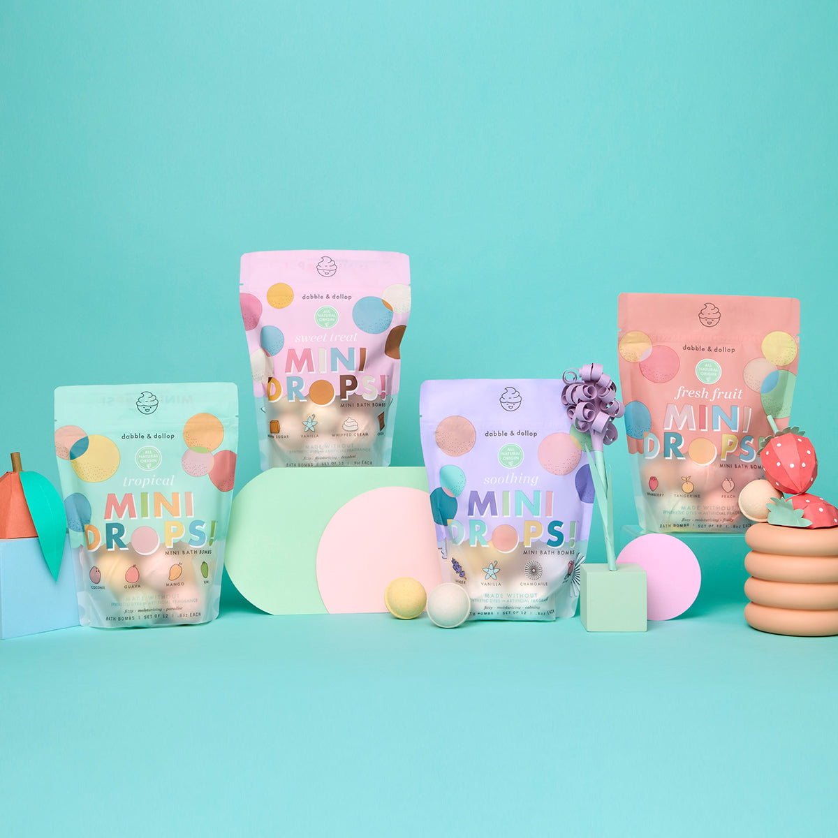 Four colorful bags of Dabble & Dollop Mini Bath Bombs are arranged among playful pastel props and shapes against a turquoise background. The packaging, reminiscent of kids’ bath bombs, features polka dots in various pastel colors for an extra burst of fun.
