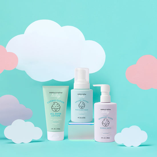 Three pastel-colored bottles of Dabble & Dollop’s Baby Bath to Bed Gift Set are displayed against a light blue background with white and pink paper clouds. This set features lotion, hair & body wash, and bubble bath—all specially formulated for sensitive baby skin.
