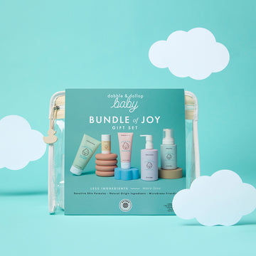 The Bundle of Joy Gift Set by Dabble & Dollop is shown in a clear pouch against a blue background with white paper clouds and features pastel-colored baby skincare products.