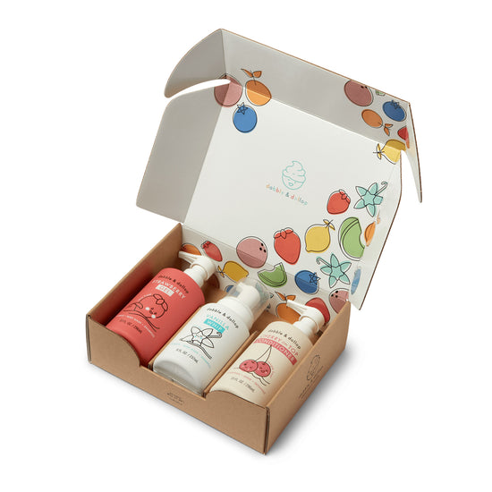 A cardboard box opens to reveal three pastel-colored bottles of Dabble & Dollop bath products, including the Cherry Berry Conditioner & Bath Bundle, with playful fruit illustrations on the packaging and inside the box lid.
