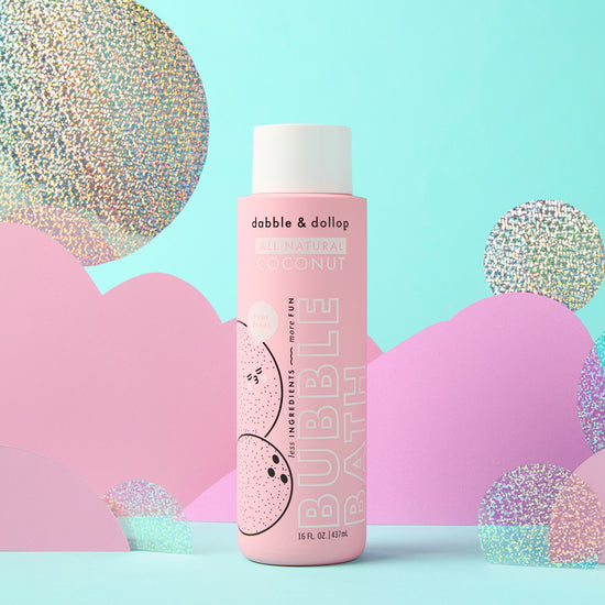 A pink bottle of Dabble & Dollop Coconut Bubble Bath sits against a pastel backdrop with abstract hills and holographic circles, featuring playful coconut graphics and bold white text—perfect for a tropical bath.