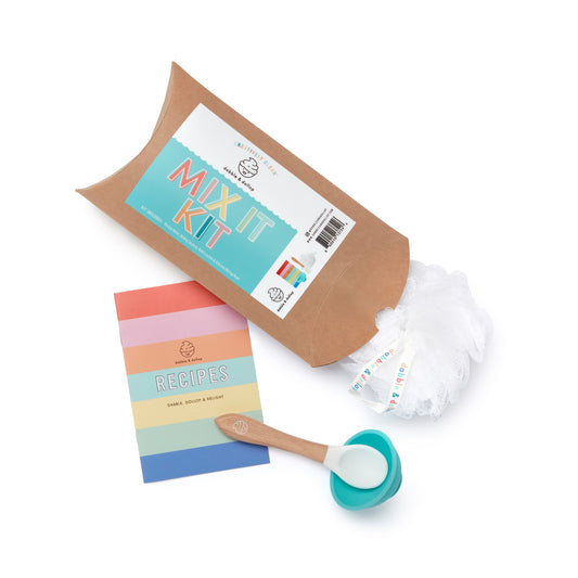 A Dabble & Dollop Mix It Kit is shown on a white background with a brown paper package, white mesh net, pastel recipe booklet, and a wooden spoon featuring a blue silicone scoop.