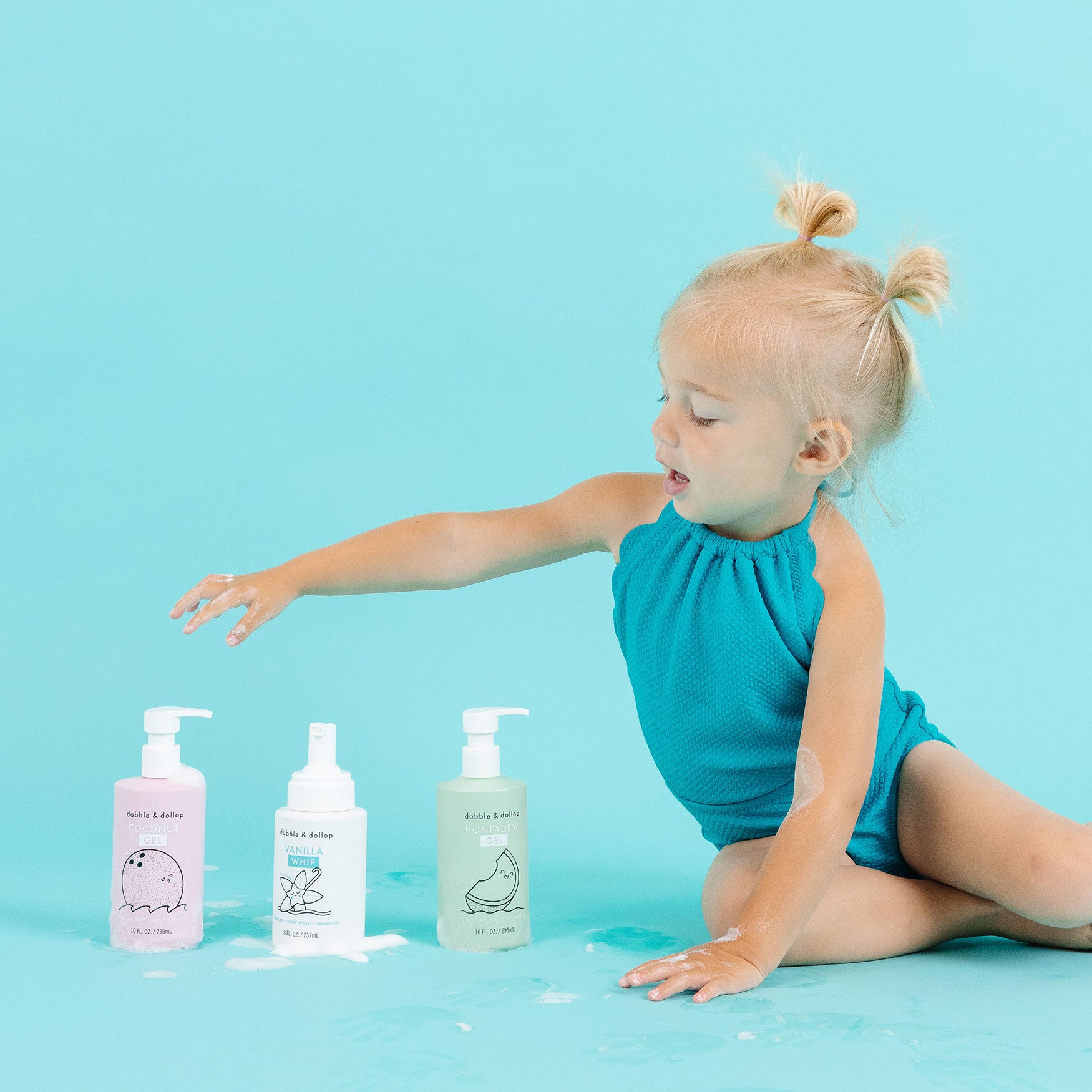 A young child in a teal romper sits on the floor, reaching for one of three colorful Dabble & Dollop A Day at the Beach Bath Bundle bottles, made with vegan and cruelty-free ingredients, surrounded by spilled foam on a teal background.