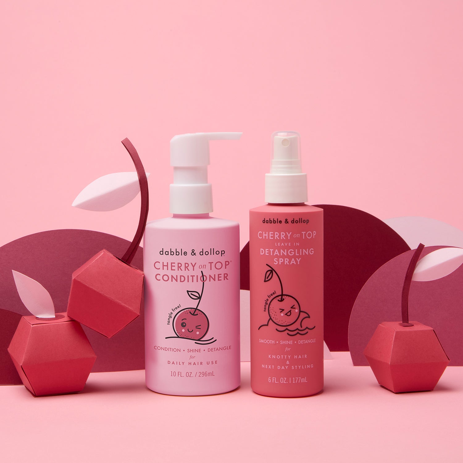 Two pink bottles from the Dabble & Dollop Cherry on Top® Detangling Duo Bundle sit among cherry decorations, featuring a kids’ conditioner and detangling spray—both with fun cherry art—to help keep hair tangle-free.