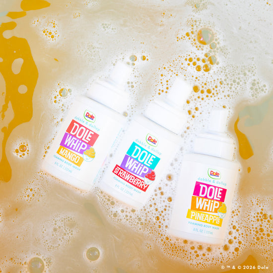 Three bottles of Dabble & Dollop Dole Whip® Foaming Body Wash Bundle in mango, strawberry, and pineapple scents float on bubbly yellow-orange water—colorful labels and fruit illustrations make for a fun, hypoallergenic body wash experience.