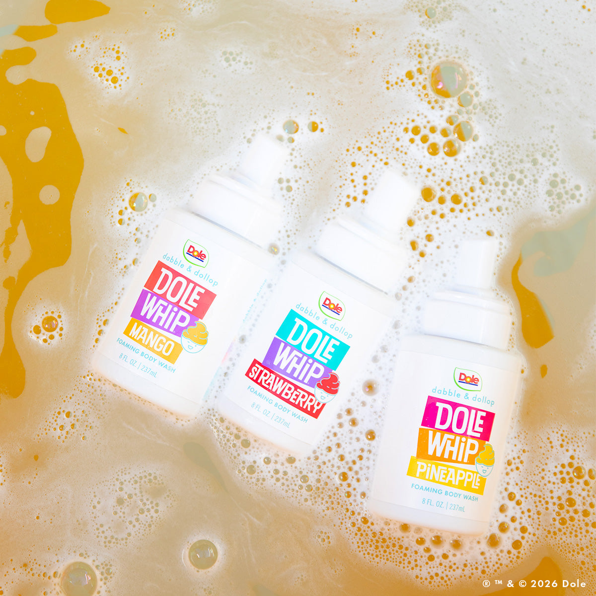 Three bottles of Dabble & Dollop Dole Whip® Foaming Body Wash Bundle in mango, strawberry, and pineapple scents float on bubbly yellow-orange water—colorful labels and fruit illustrations make for a fun, hypoallergenic body wash experience.