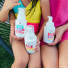 A child in a yellow swimsuit and an adult in a pink dress sit together, each holding Dabble & Dollop's Dole Whip® Pineapple Foaming Body Wash with natural origin fragrance.