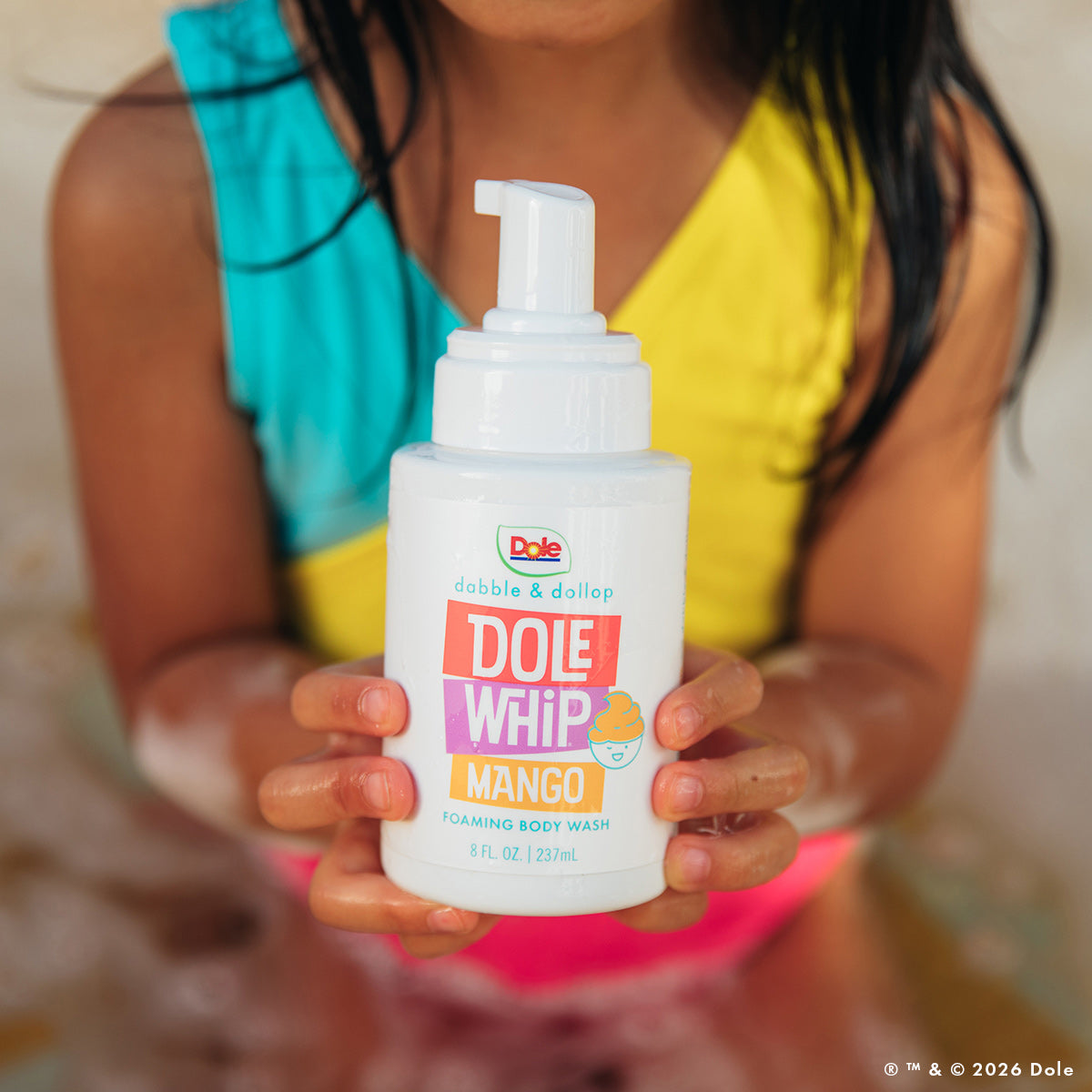 A child in a colorful swimsuit holds a bottle of Dabble & Dollop Dole Whip® Mango Foaming Body Wash, surrounded by bubbles and water, enjoying its playful natural mango fragrance.