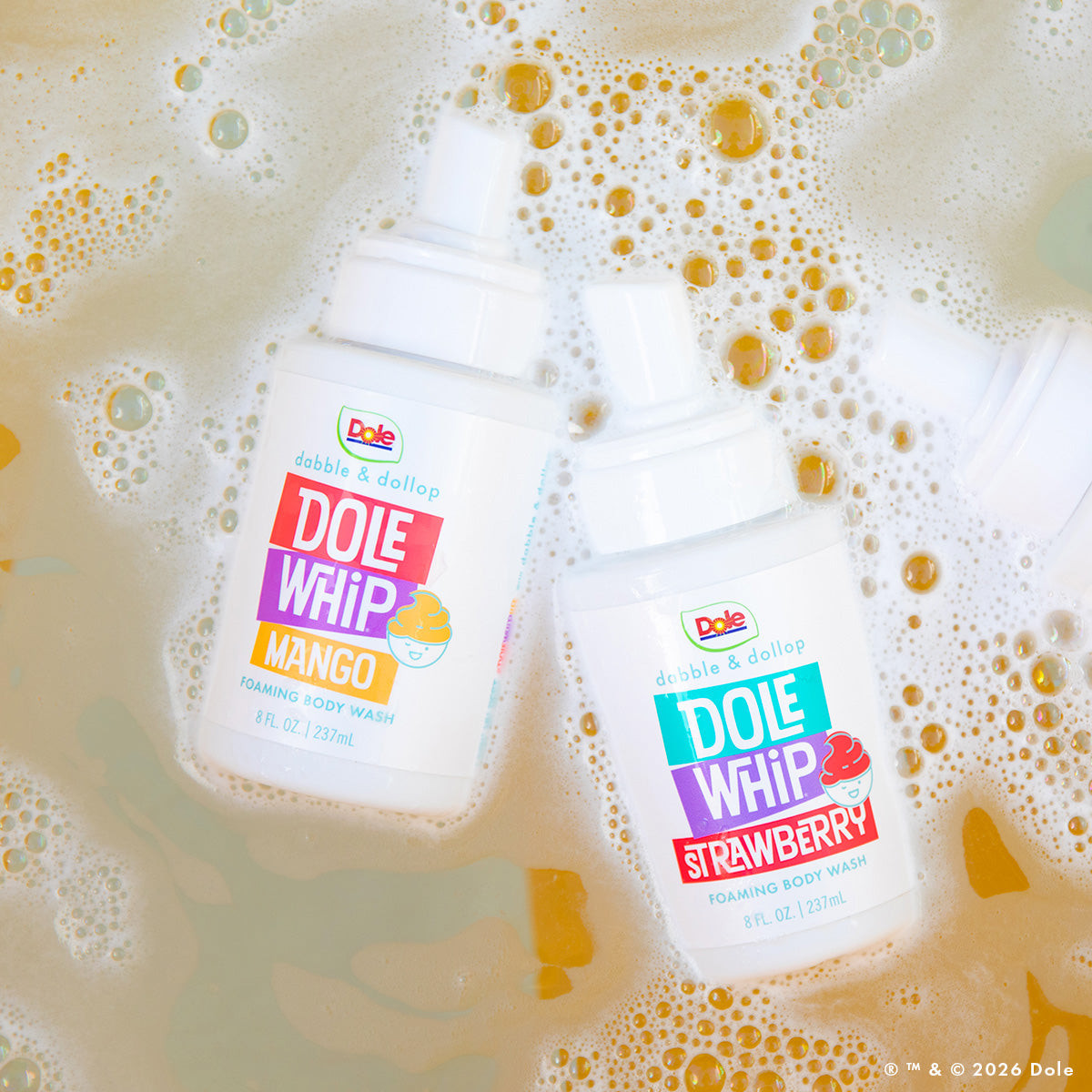 Two bottles of Dabble & Dollop Dole Whip® Mango Foaming Body Wash, in mango and strawberry scents, float on bubbly soapy water. Their vibrant labels and fruity fragrances stand out against the light background.