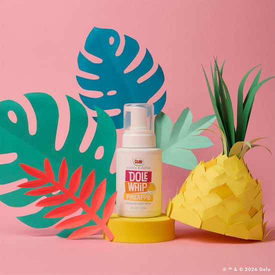 A bottle of Dabble & Dollop Dole Whip® Pineapple Foaming Body Wash sits on a pink background, surrounded by colorful paper leaves and a paper pineapple.